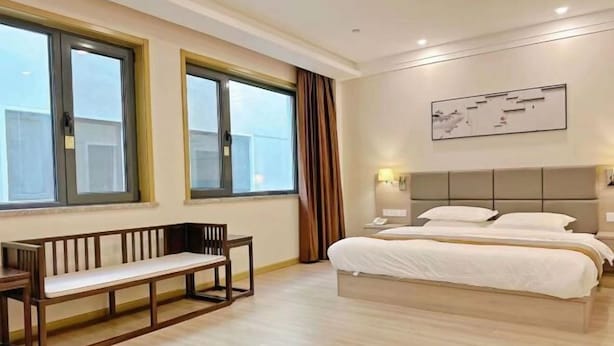 A hotel room with a bed, a bench, a bedside table, a lamp, and a window with curtains. - Hancun River Villa (Beijing)