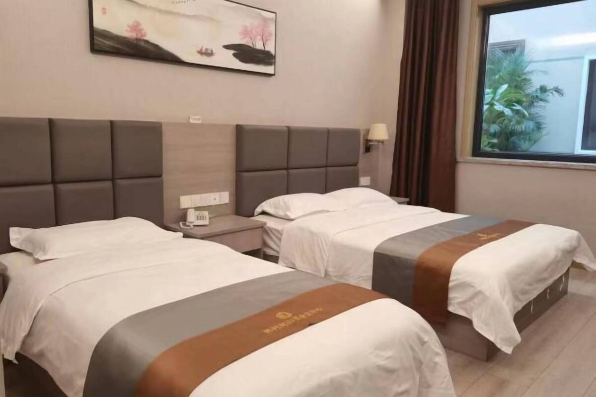 A hotel room with two beds, a nightstand, a painting on the wall, and a window with a view of greenery.