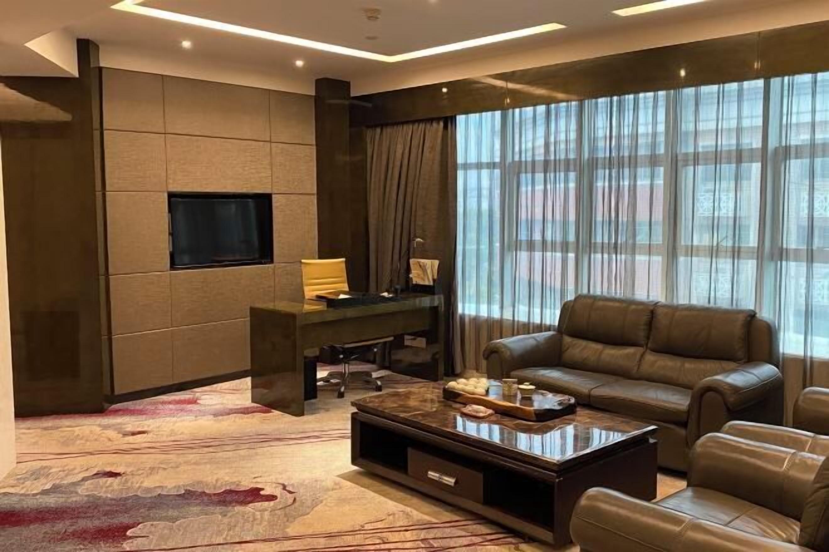 A modern living room with a leather sofa, a coffee table, a TV mounted on the wall, and a large window with curtains.