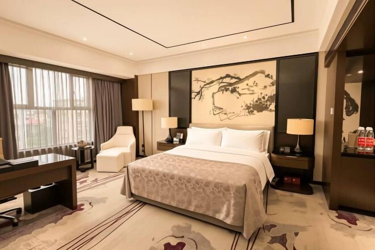 Ein Hotelzimmer mit Bett, Schreibtisch, Stuhl, zwei Nachttischen mit Lampen und einem großen Kunstwerk an der Wand.