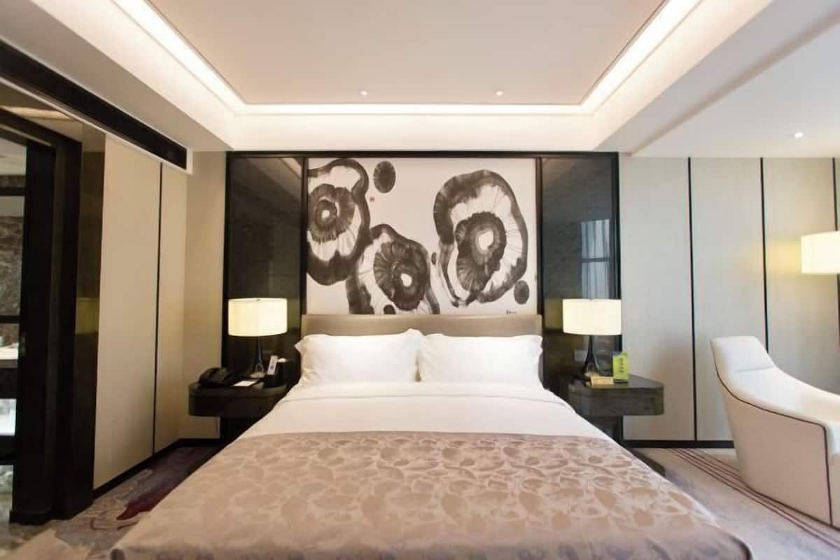 Ein modernes Hotelzimmer mit einem großen Bett, zwei Nachttischen mit Lampen, einem großen abstrakten Kunstwerk an der Wand und einem weißen Sessel.