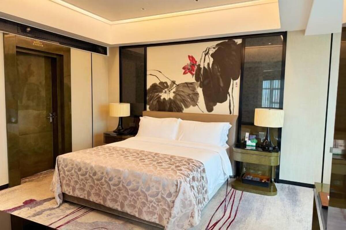 Ein Hotelzimmer mit einem Bett, zwei Nachttischlampen, einem Nachttisch, einem großen Wandbild und einer Tür.