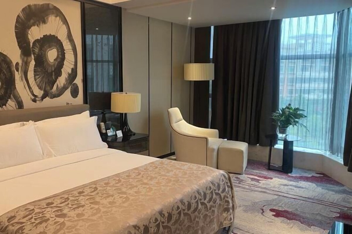 A hotel room with a large bed, a desk with a lamp, a chair, and a window with curtains.