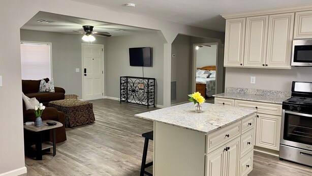A modern kitchen with white cabinetry, a granite countertop, and a stainless steel stove. There is a dining area with a brown sofa and a small table. - Twin Oaks Cozy Getaway in Lake Charles (Lake Charles)