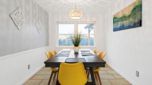 A dining area with a rectangular table, yellow chairs, a hanging light fixture, and a window with a view. - Near Highways, Family Friendly, Garage, Foosball, Grill, Dart, Tesla Charger (Plano)