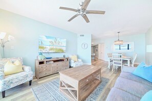 A living room with a ceiling fan, a television, a dining area with a table and chairs, and a light blue wall.
