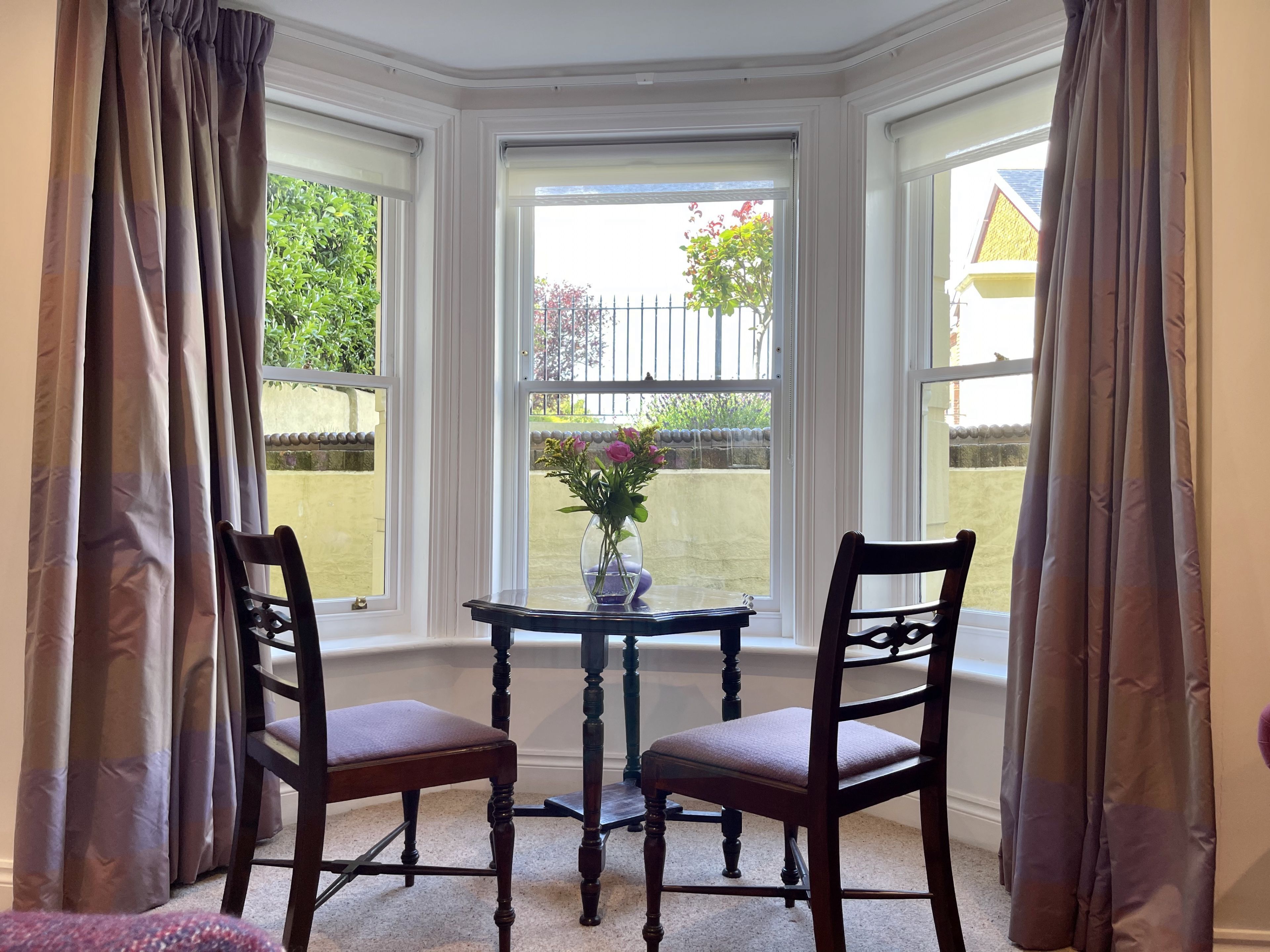 A bay window with two chairs and a table, a vase with flowers, and a view of a garden and fence.