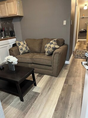 A living room with a brown sofa, two patterned pillows, a small table with a vase, and a kitchen area with cabinets and utensils.