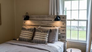 Ein Schlafzimmer mit einem hölzernen Kopfteil, zwei Nachttischlampen, einem Bett mit gemusterten Kissen und einem Fenster mit Vorhängen.