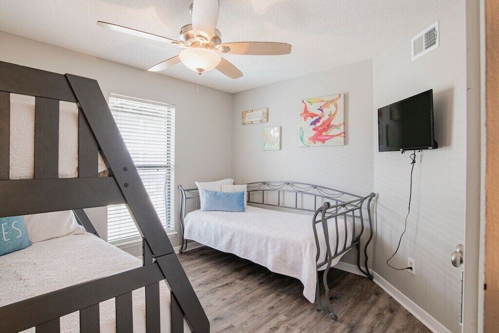 Un dormitorio con literas, un ventilador de techo, una cama con edredón blanco, un televisor montado en la pared y obras de arte decorando la pared.