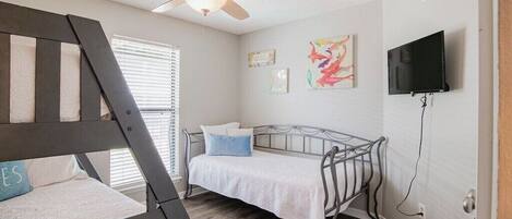 Un dormitorio con literas, un ventilador de techo, una cama con edredón blanco, un televisor montado en la pared y obras de arte decorando la pared.