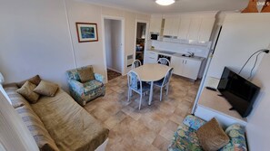 Ein gemütlicher Wohnraum mit Sofa, einem gemusterten Sessel, einem Esstisch mit Stühlen, einer Küchenzeile mit Schränken und Geräten sowie einem Fernseher.