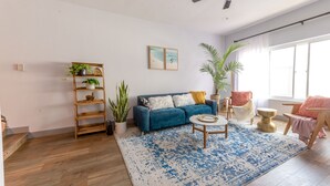Un salon avec un canapé bleu, une étagère en bois en forme d’échelle ornée de plantes, un tapis à motifs et une grande fenêtre.