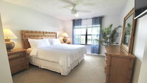 Ein Schlafzimmer mit einem Bett, Nachttischen, einer Kommode, einem Deckenventilator, einem Fenster mit Vorhängen und einer Topfpflanze.