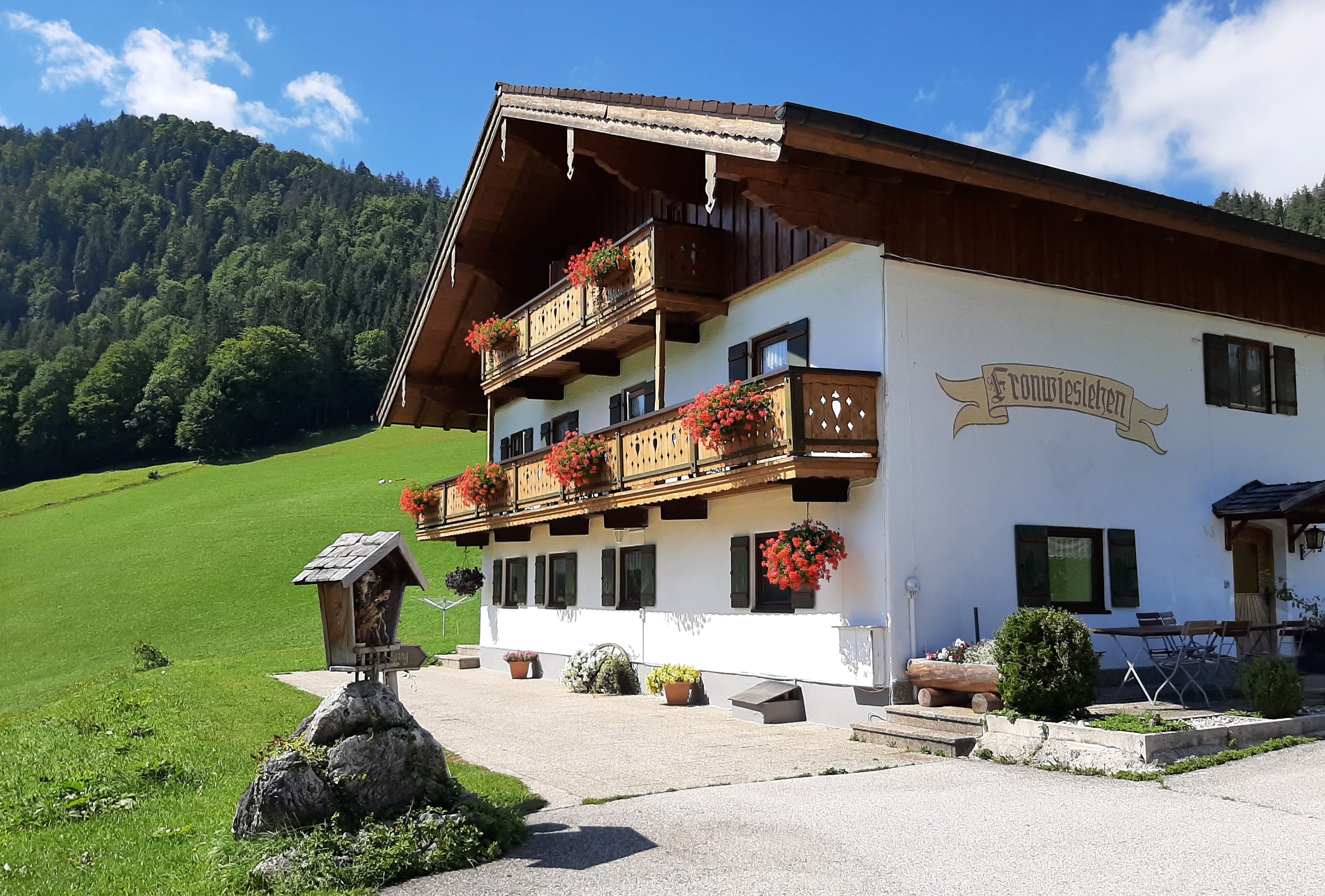A traditional Alpine-style building with a balcony adorned with flowers, set against a backdrop of a green hillside and a clear sky.