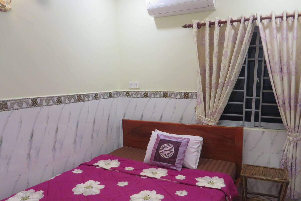 Un dormitorio con cama, ventana con cortinas, un taburete de madera y aire acondicionado de pared.