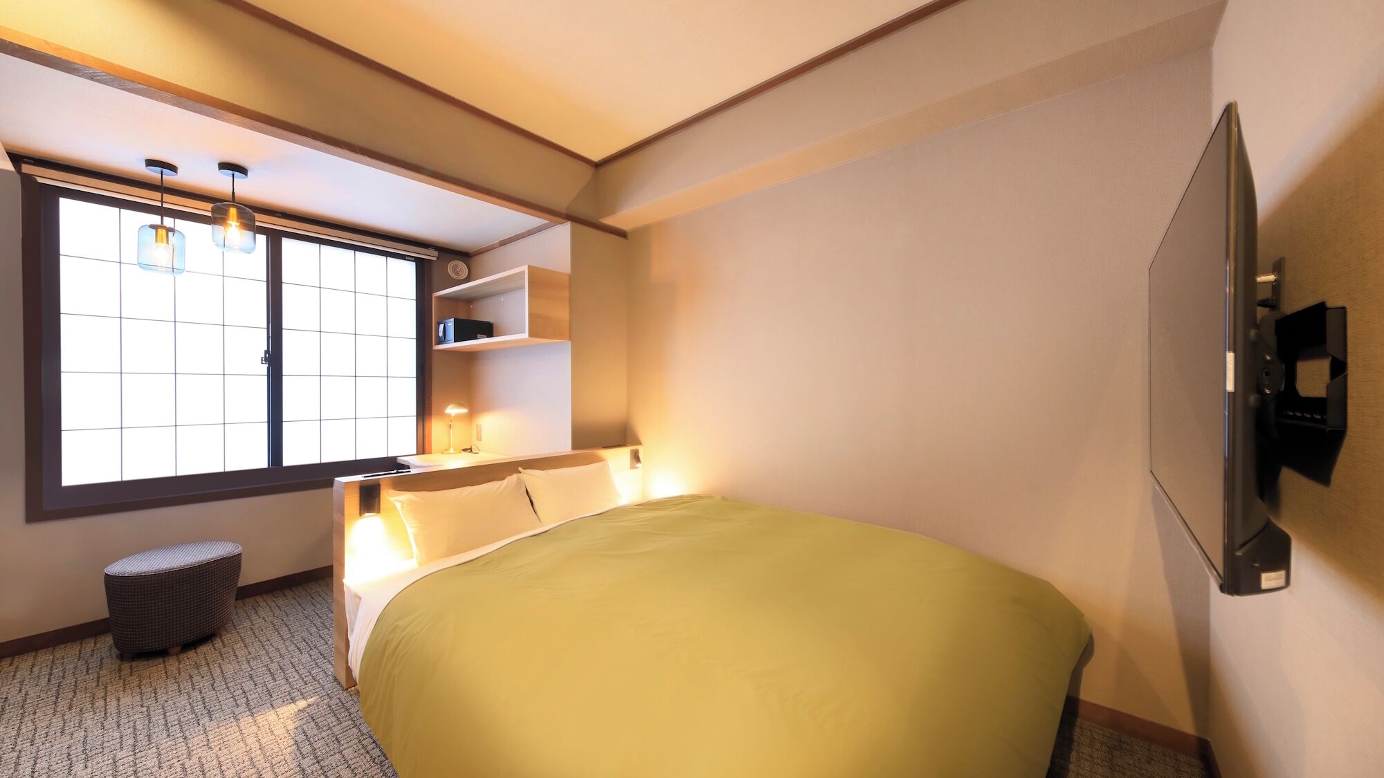 Une chambre d’hôtel avec un lit, un téléviseur fixé au mur, une fenêtre avec des stores, une table de chevet et un petit tabouret.