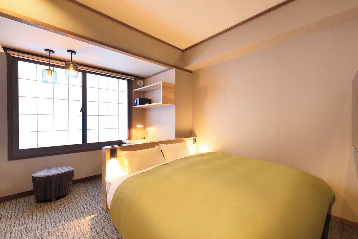 A hotel room with a bed, a TV mounted on the wall, a window with blinds, a bedside table, and a small stool.