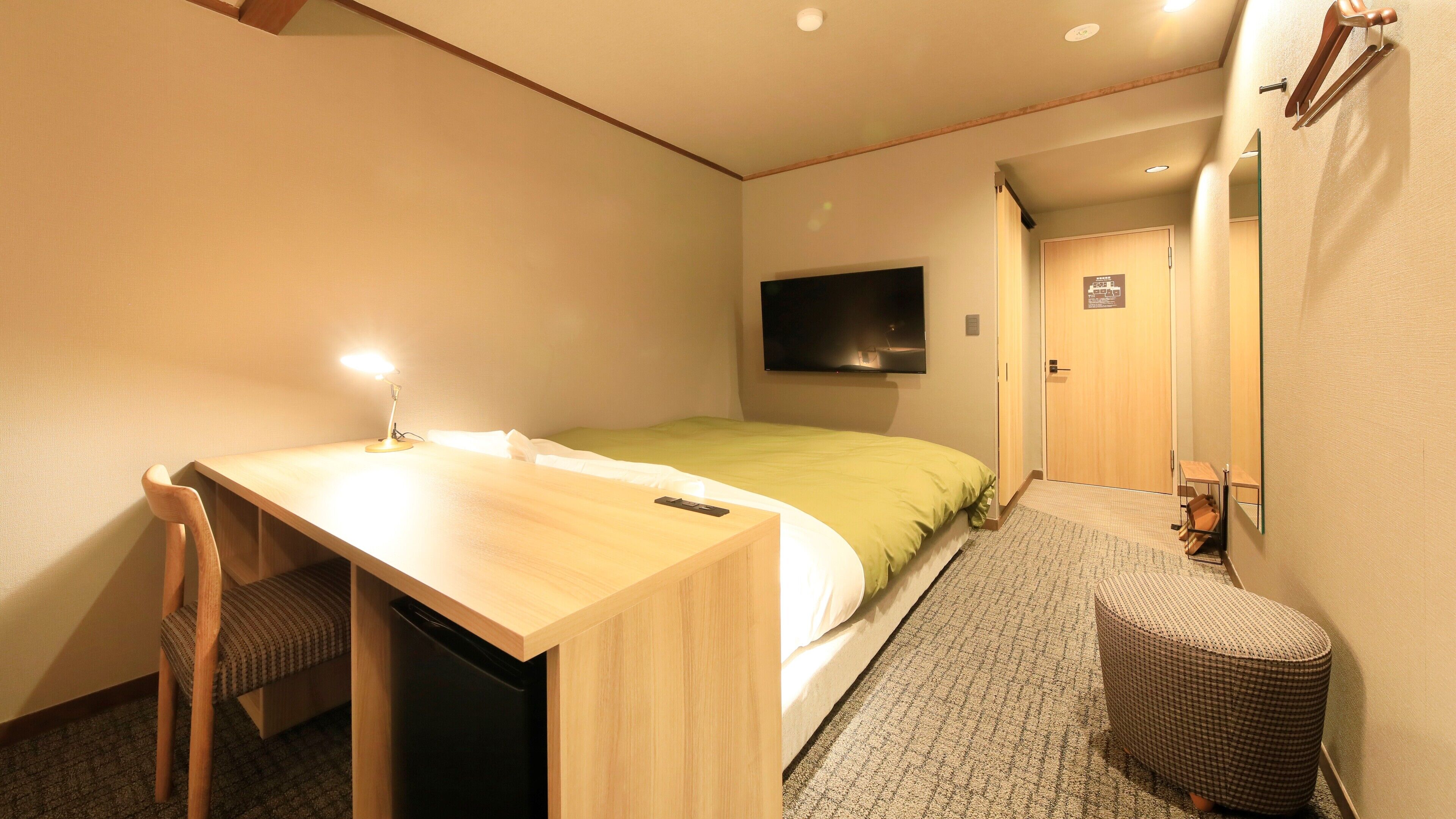 A compact hotel room with a bed, a desk with a chair, a television mounted on the wall, and a small wicker stool.