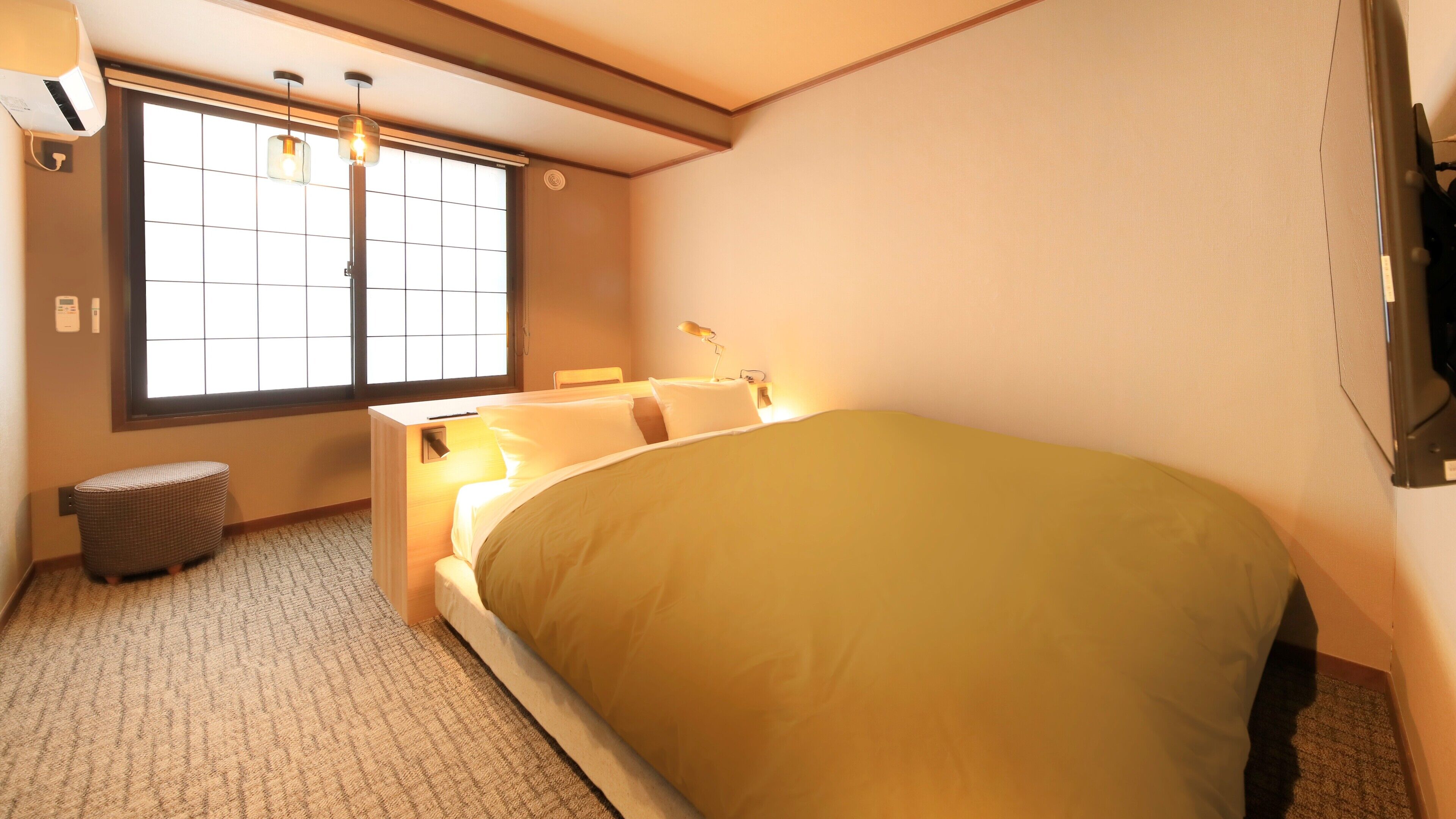 A hotel room with a bed, bedside table, lamp, window, and a small stool.