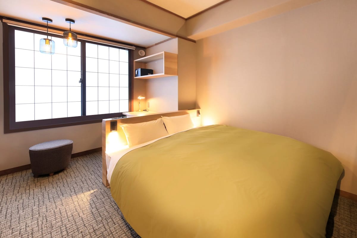 A hotel room with a large bed, a window with blinds, a bedside table with a lamp, a small seating area, and a TV mounted on the wall.