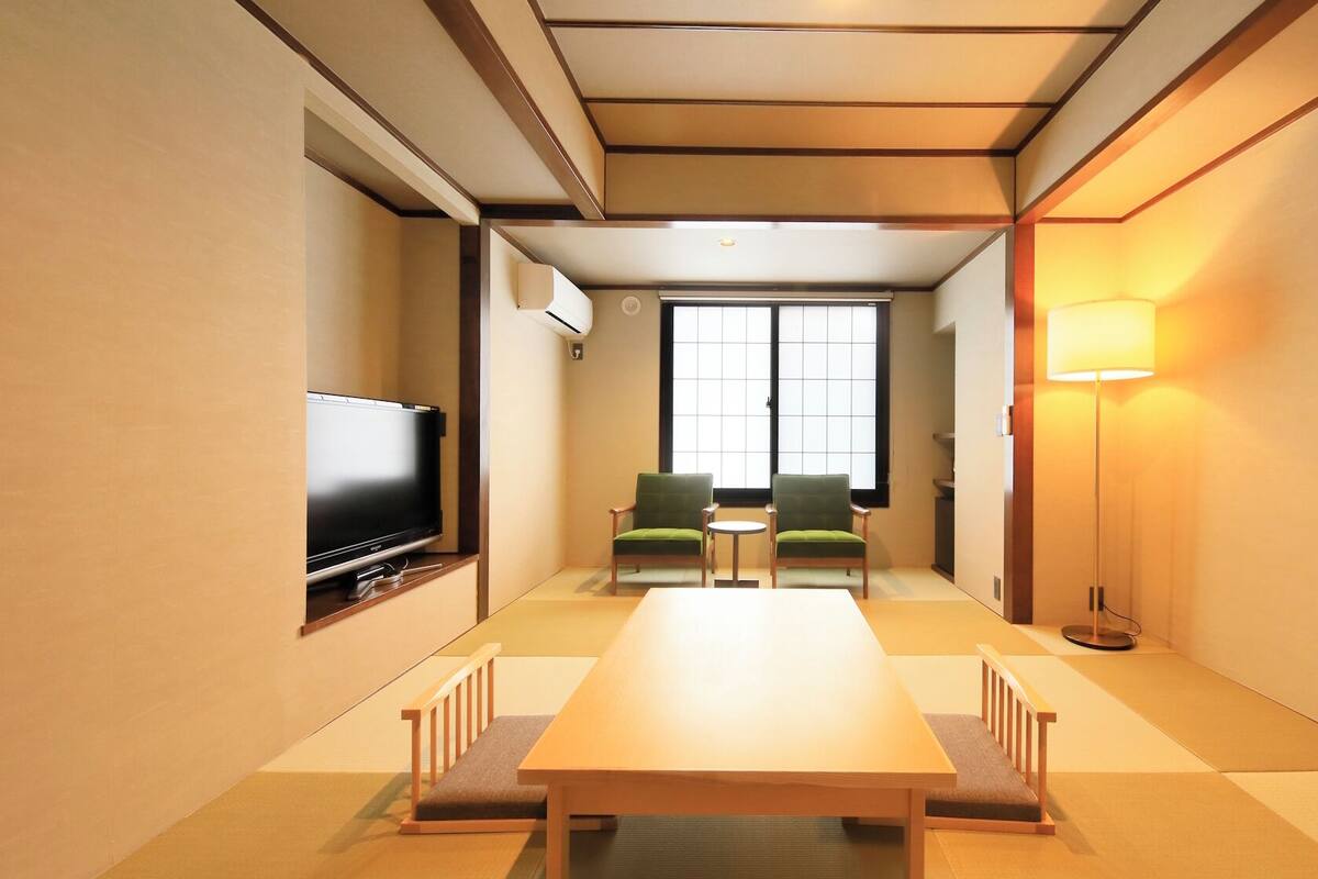 A hotel room with a TV, a wooden table, two chairs, a small table, and a floor lamp.