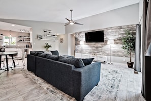 A modern living room with a dark gray sofa, a dining area with a table and chairs, a kitchen in the background, a mounted TV, and a ceiling fan.