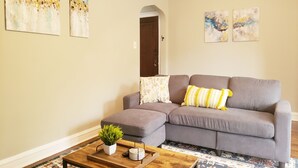 A living room with a grey sectional sofa, a wooden coffee table with a plant, and a patterned rug. There are two abstract paintings on the walls. - Comfy Bed/Free Parking&Wifi /NearHosp/ORD/MDW/DT (Berwyn)