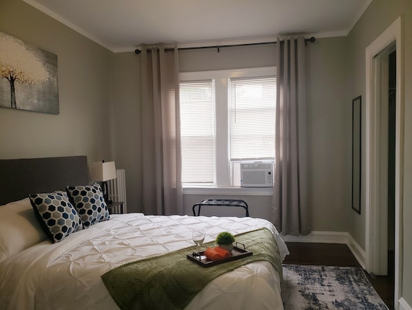 Une chambre d’hôtel avec un grand lit, une fenêtre avec des stores, un appareil de climatisation, une table de chevet avec une lampe et un tableau encadré au mur.