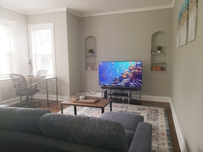 Un salon avec un canapé gris, une table basse, un téléviseur posé sur un meuble, un bureau avec une chaise et des étagères ornées d’objets décoratifs.