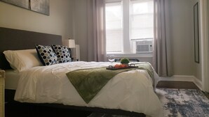 A neatly made bed with white linens and a green throw blanket, a bedside table with a glass and orange towel, and a window with blinds. - Comfy Bed/Free Parking&Wifi /NearHosp/ORD/MDW/DT (Berwyn)