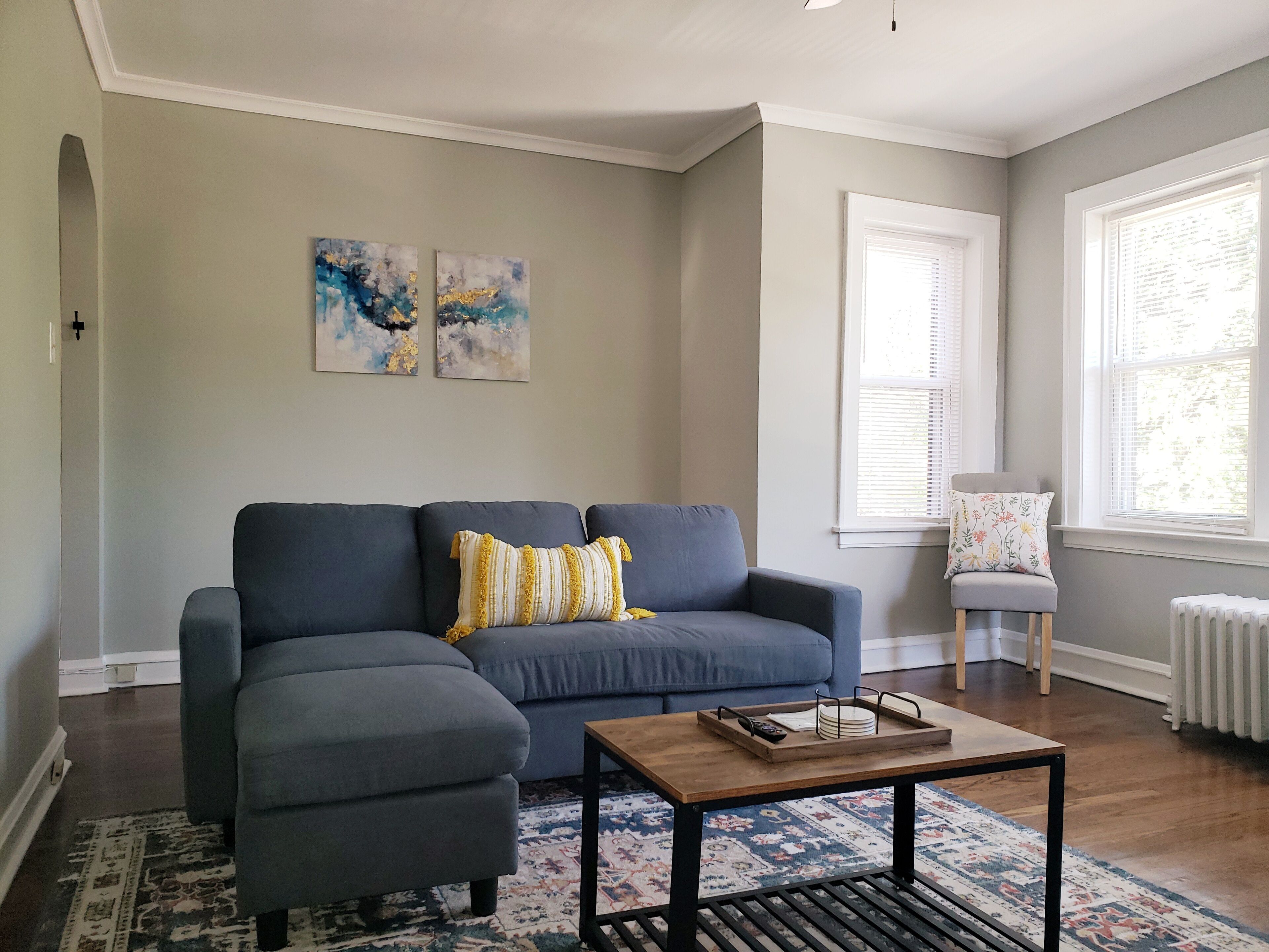 A living room with a grey sectional sofa, a wooden coffee table, and a patterned rug. There are abstract paintings on the wall and a window with blinds.