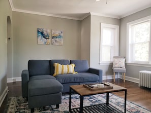 Un salon avec un canapé d’angle gris, une table basse en bois et un tapis à motifs. Les murs sont ornés de tableaux abstraits et une fenêtre est dotée de stores.
