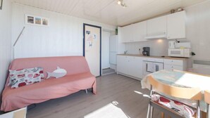Una habitación compacta con un sofá rosa, una mesa pequeña con mantel estampado y una zona de kitchenette con armarios y electrodomésticos blancos.