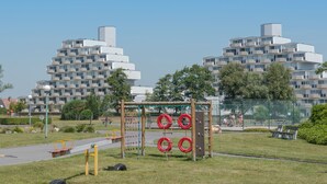 Un parque infantil con estructuras para escalar y un banco, rodeado de una cerca y árboles, con edificios modernos al fondo.