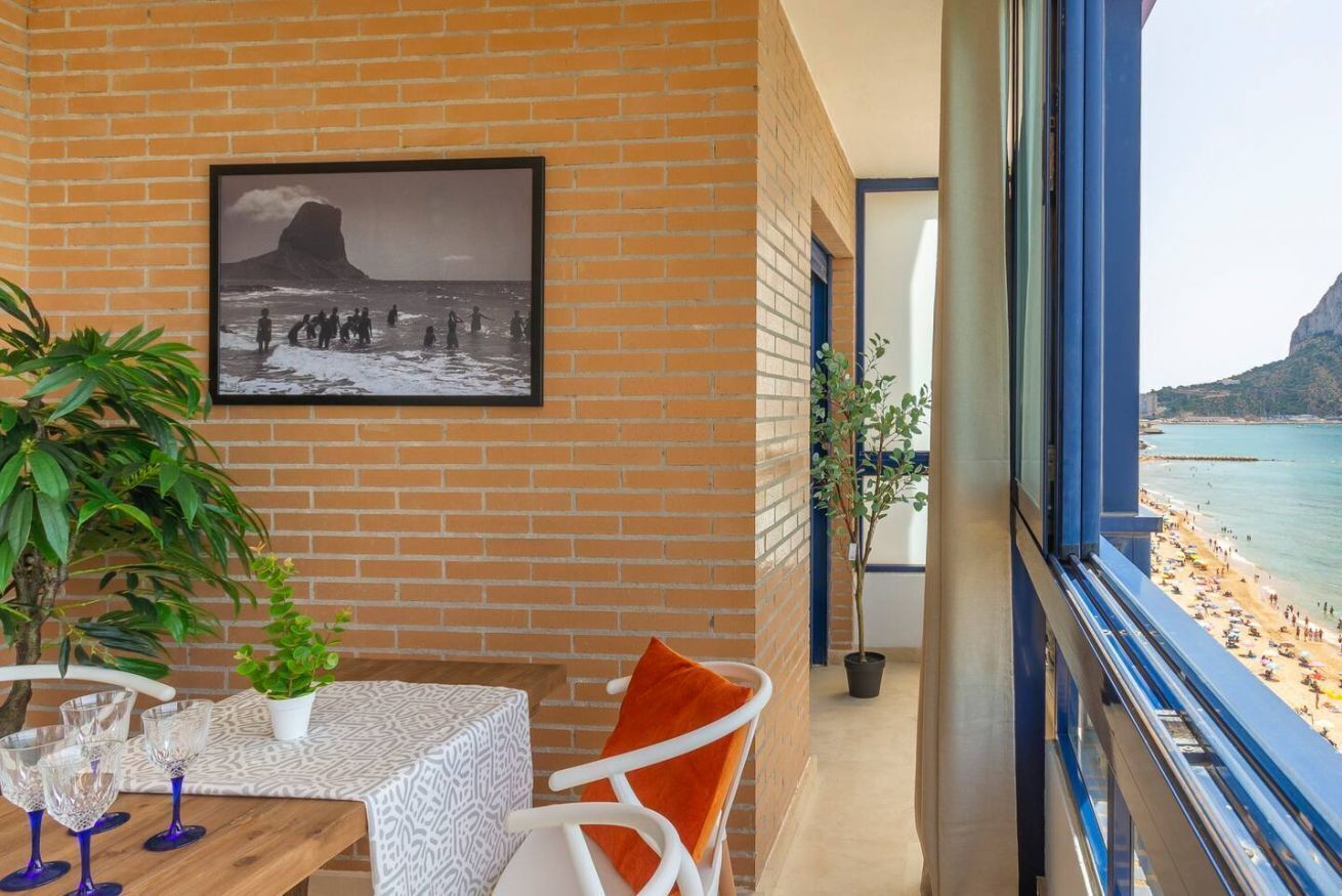 En balkon med udsigt over stranden, et bord med glas, en stol og et billede i ramme på væggen.