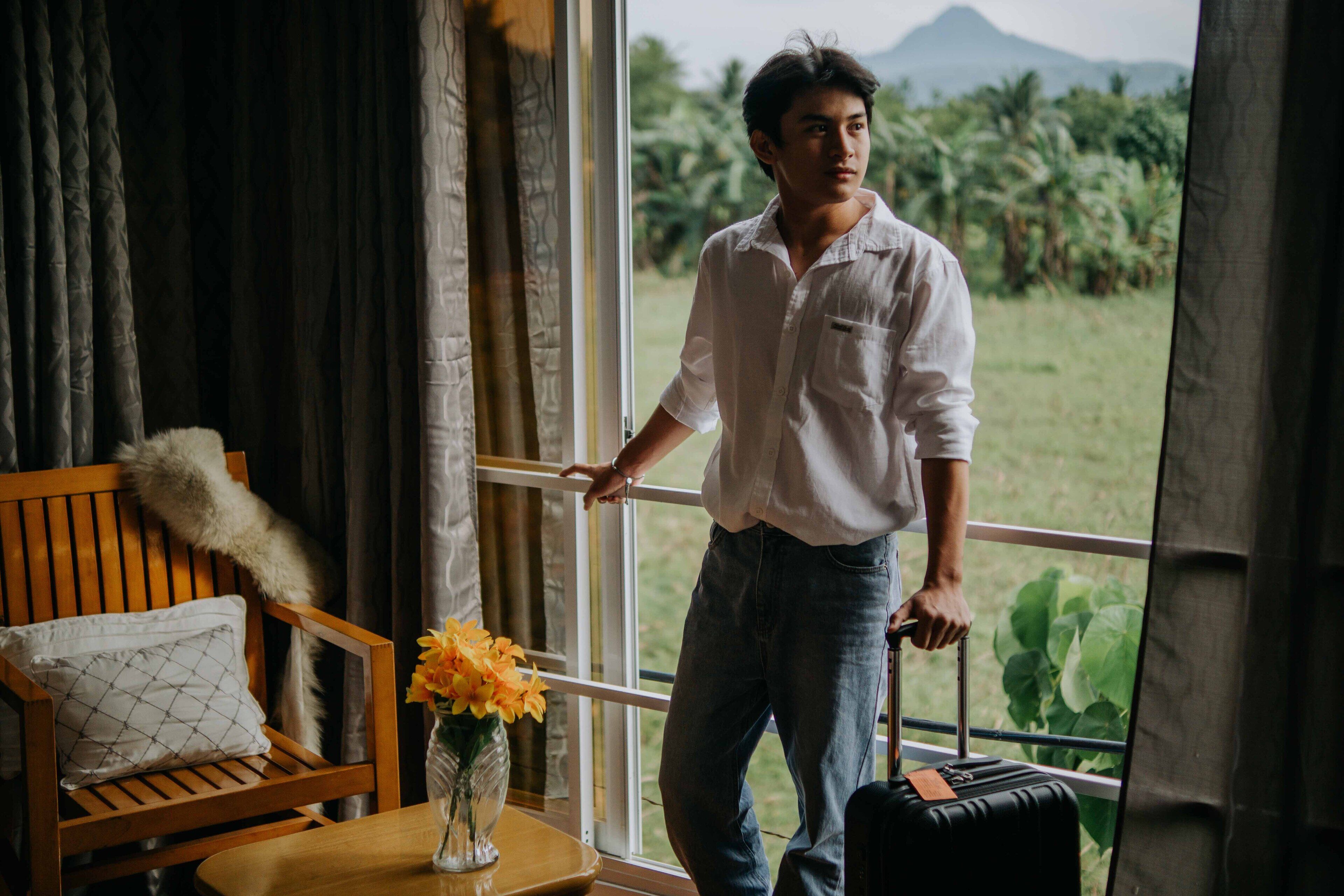 A person standing by a window with a view of greenery and a mountain, holding a suitcase.