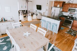 A modern kitchen with wooden cabinets, a stainless steel refrigerator, and a microwave. There is a dining area with a wooden table and white chairs. - ★ "This house is beautiful inside and out." (Nags Head)