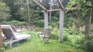 A garden with two wooden chairs and a table, surrounded by greenery and a stone wall. - Walk to the ocean, very private. Secluded, woodsy location.  (Gloucester)