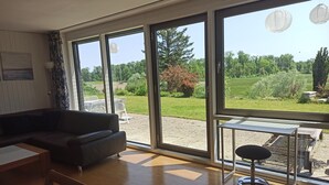 Ein modernes Wohnzimmer mit einem dunkelgrauen Sofa, einem Holztisch und einem kleinen runden Tisch mit Barhocker. Große Fenster bieten Blick auf einen grünen Garten und einen blauen Himmel.