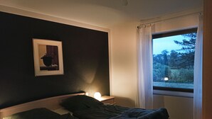 Ein Schlafzimmer mit Bett, Nachttischlampe, einem gerahmten Bild an der Wand und einem Fenster mit Blick auf Bäume.