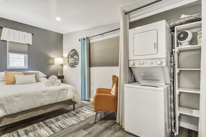 A bedroom with a bed, a chair, a washing machine, and a shelf with a fan.