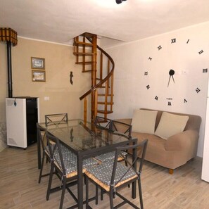 A dining area with a glass table, four chairs, a spiral staircase, a refrigerator, a sofa, and a wall clock.