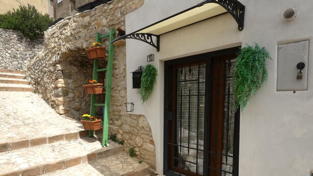 A stone staircase leading to a building with a window and a door. There are potted plants on a green metal structure and hanging from the wall. - Escape To Umbria, Little Alley (Porchiano del Monte)