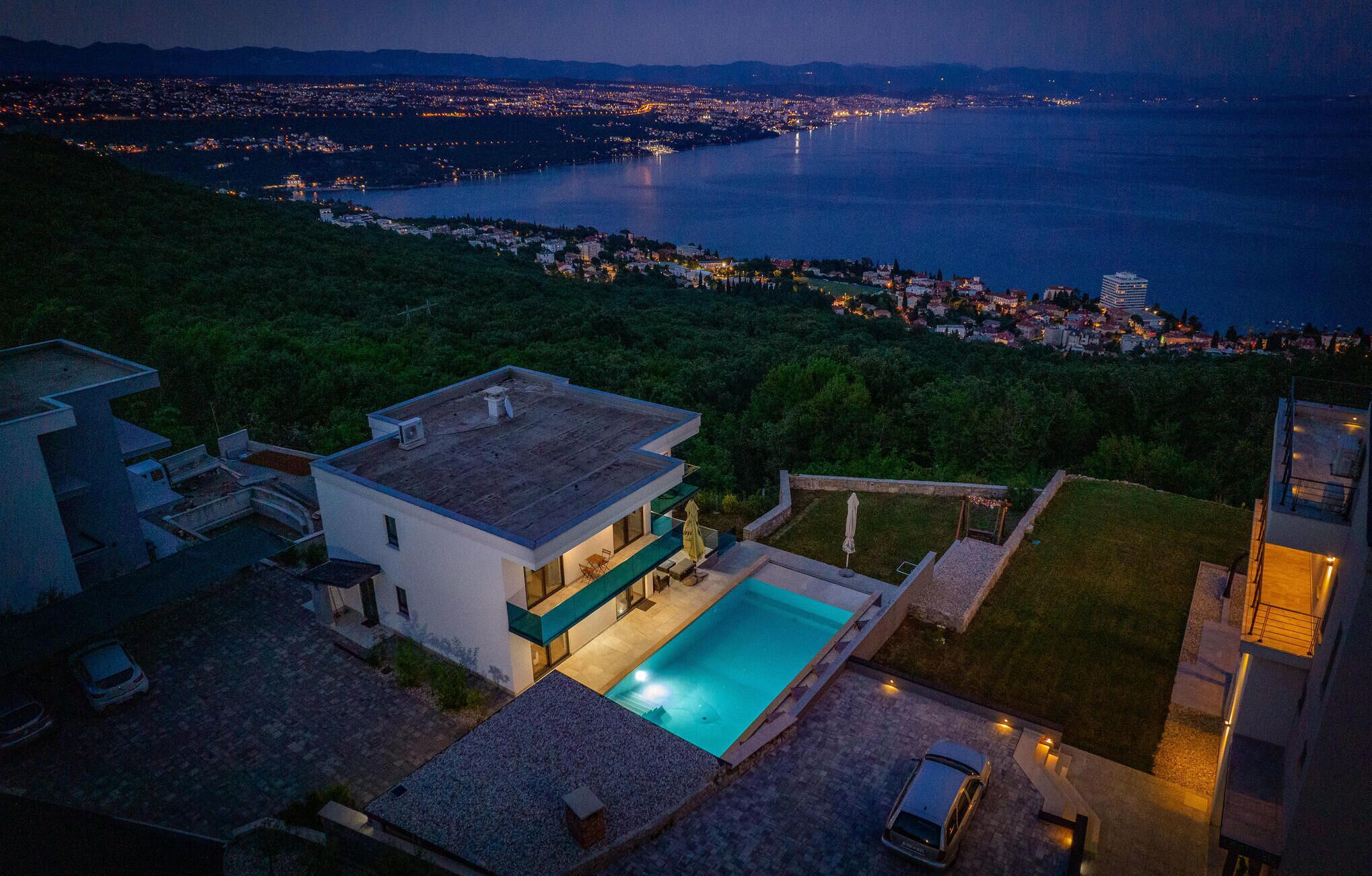 Ein modernes Haus mit Swimmingpool, umgeben von üppiger Vegetation und mit Blick auf eine beleuchtete Küstenstadt bei Nacht.