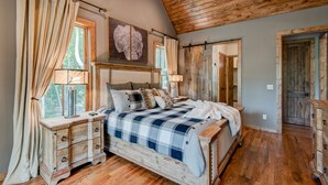 Een slaapkamer met een houten plafond, een groot bed met een geruit sprei, een houten dressoir en een raam met gordijnen.