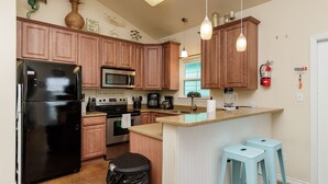 A kitchen with wooden cabinets, a black refrigerator, a microwave, a stove, and a sink. There are two light blue stools and a trash can.