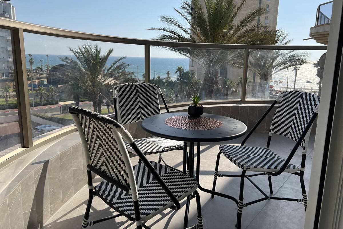 Un balcon avec une table ronde et quatre chaises, donnant sur un paysage urbain ponctué de palmiers et offrant une vue sur la mer.