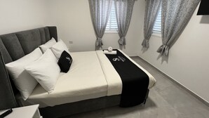 A bedroom with a bed, pillows, a blanket, curtains, an air conditioning unit, and a bedside table. - Masarik Apartments (Bat Yam)