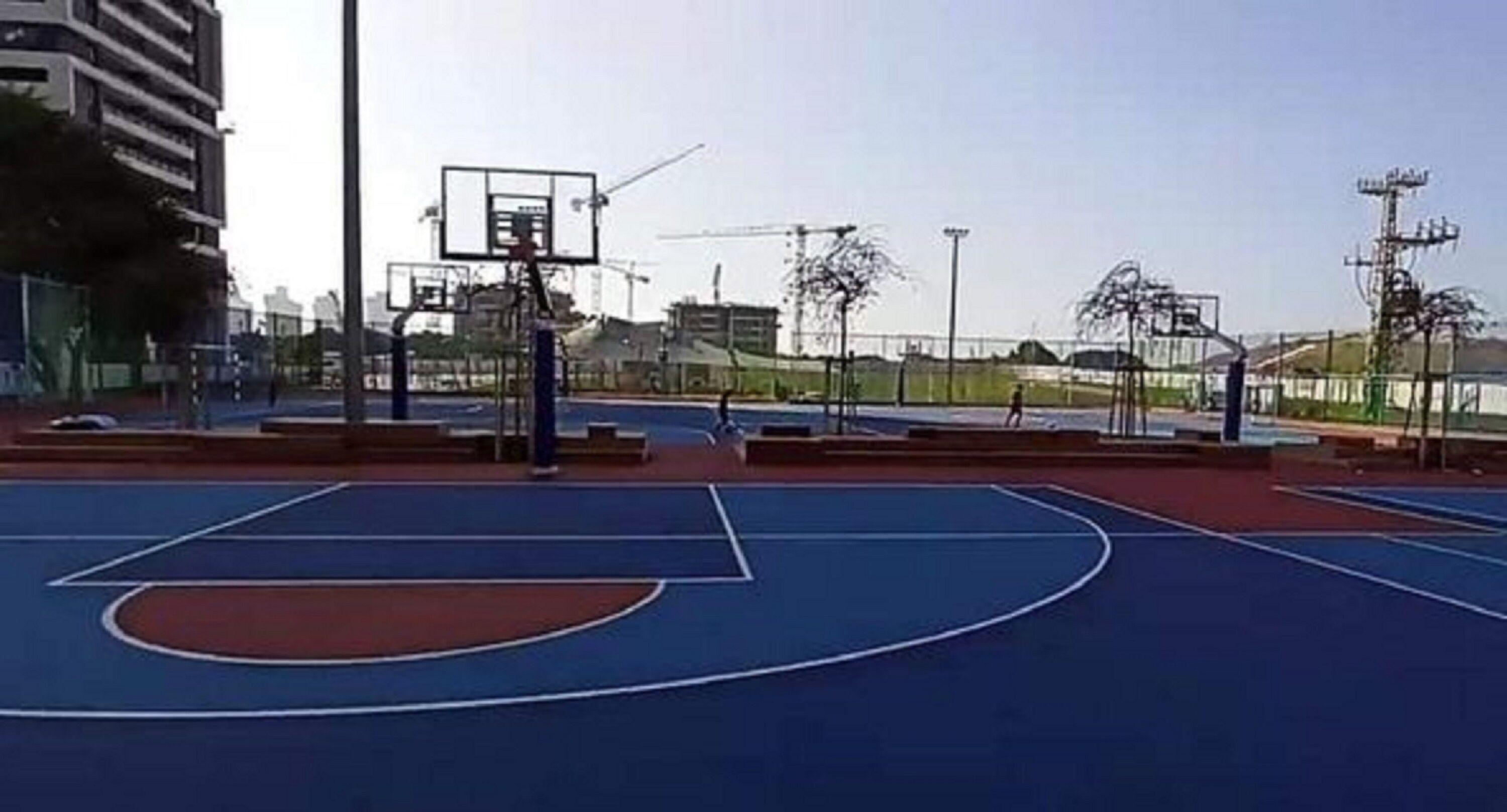 En udendørs basketballbane med blå spilleflade, en basketballkurv og et hegn.